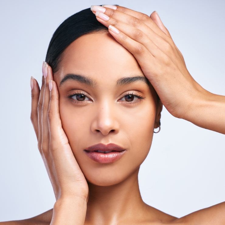 Person with natural makeup touching their face with both hands against a plain background.