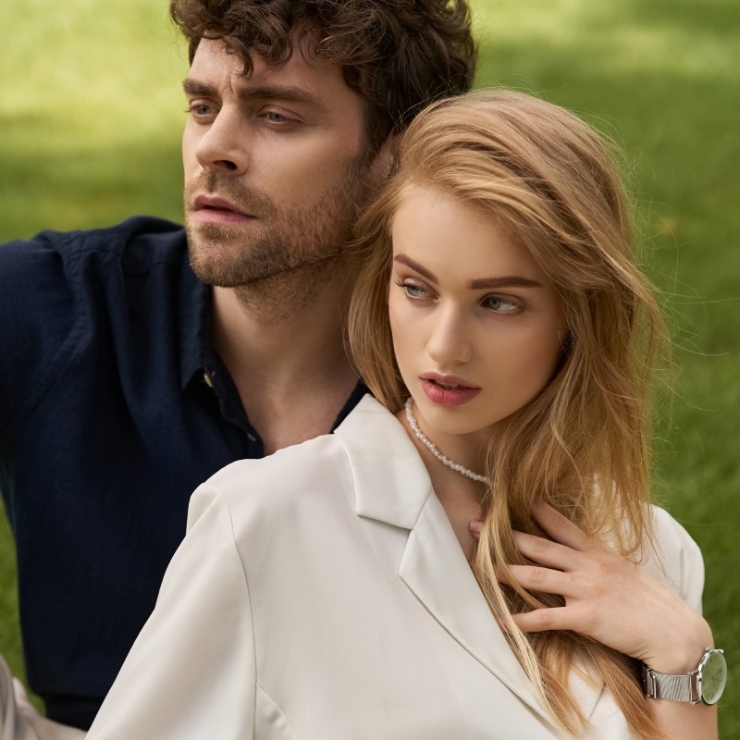 A man and woman sitting on grass, looking away. The woman has blonde hair and is wearing a white top and a necklace. The man has curly hair and is wearing a dark shirt.