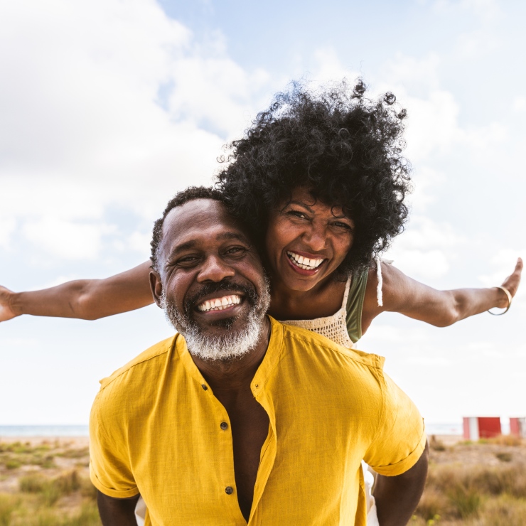 A joyful couple stands on a beach, with the man giving the woman a piggyback ride. They are both smiling broadly, and the sky is clear with a few clouds.