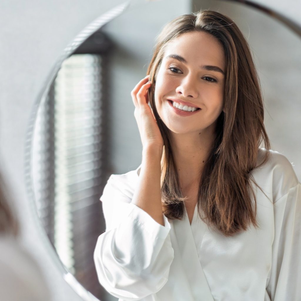 Woman smiling in front of a round mirror, wearing a white top, with one hand touching her face.