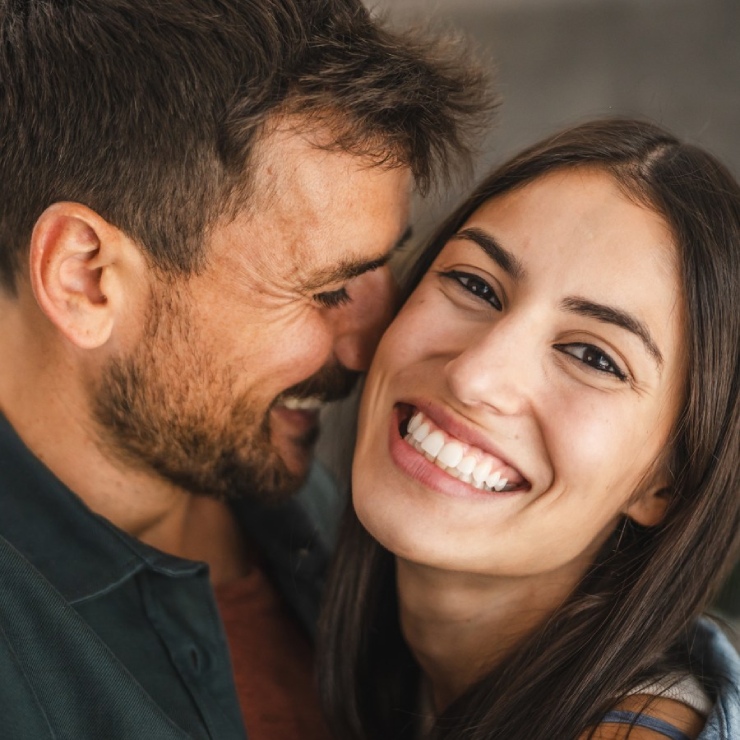 A couple smiling and embracing closely, with the woman facing the camera and the man looking at her. Their expressions are happy and relaxed.