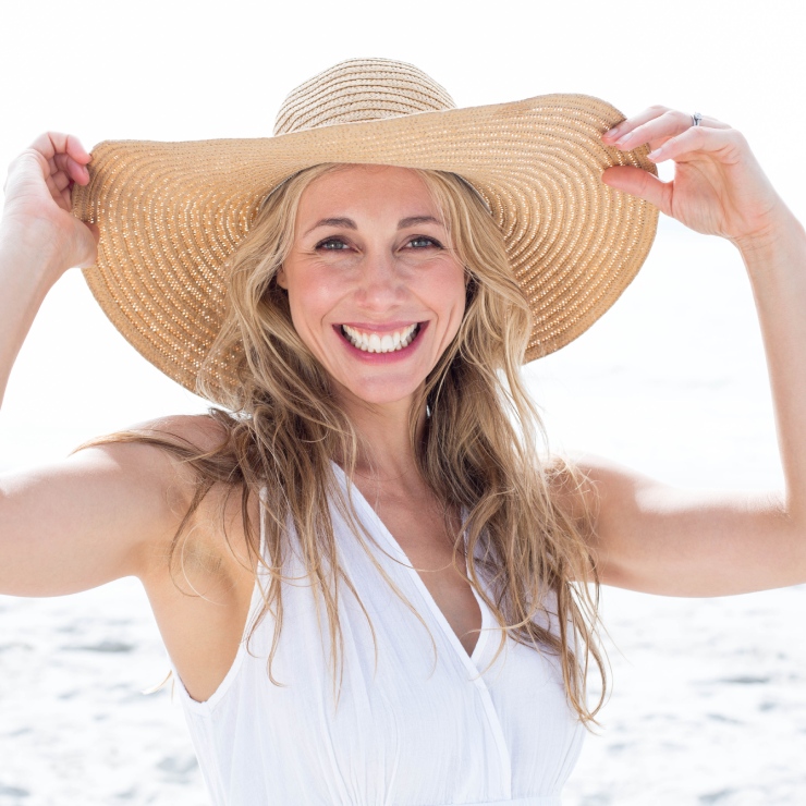 A woman with long hair smiles broadly, wearing a wide-brimmed straw hat and a sleeveless white top, standing on a beach.