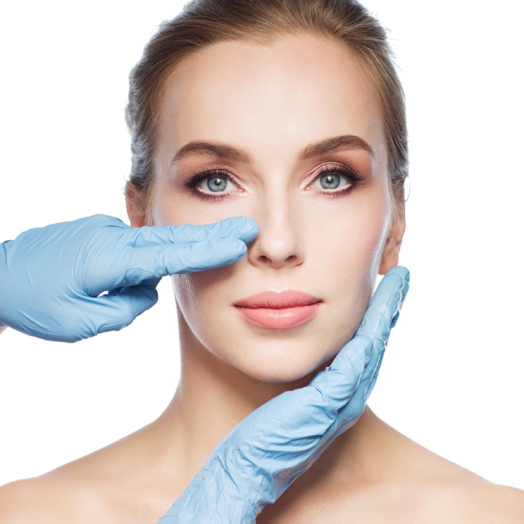 A woman with blue eyes is being examined by hands in blue gloves, one hand touching her cheek and the other adjusting her nose, against a white background.