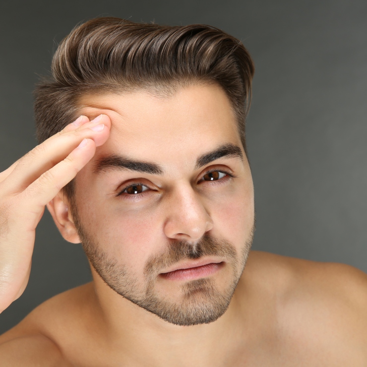 A man with short, brown hair touches his forehead while looking at the camera. He has a neutral expression and a trimmed beard. The background is gray.