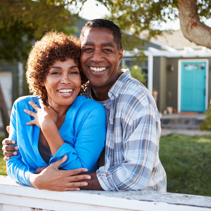 A couple smiles while leaning on a white fence in front of a house with a blue door, surrounded by trees.