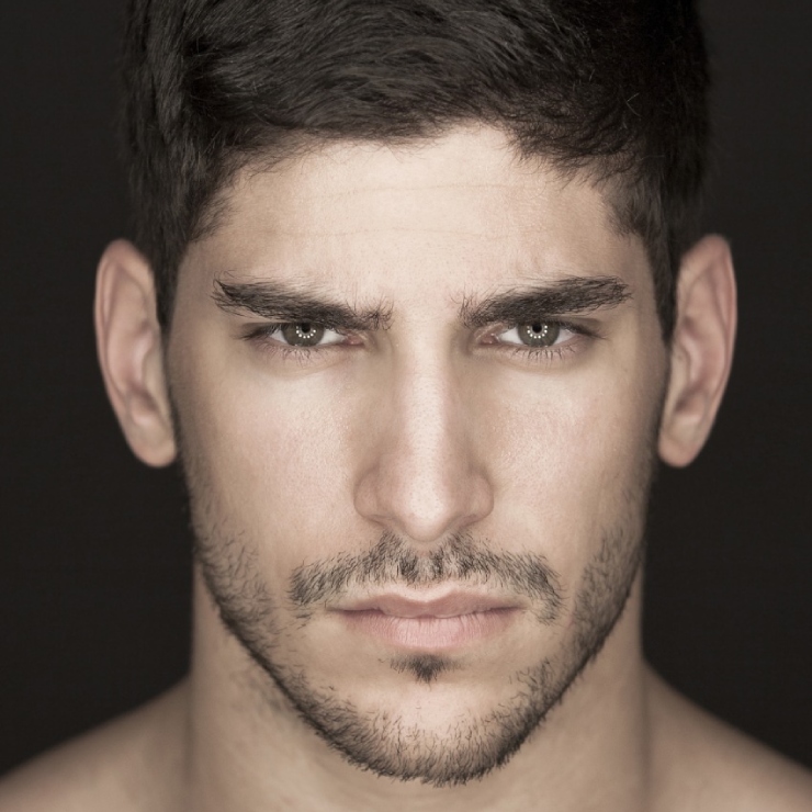 A close-up of a person with short dark hair, a beard, and a serious expression against a dark background.