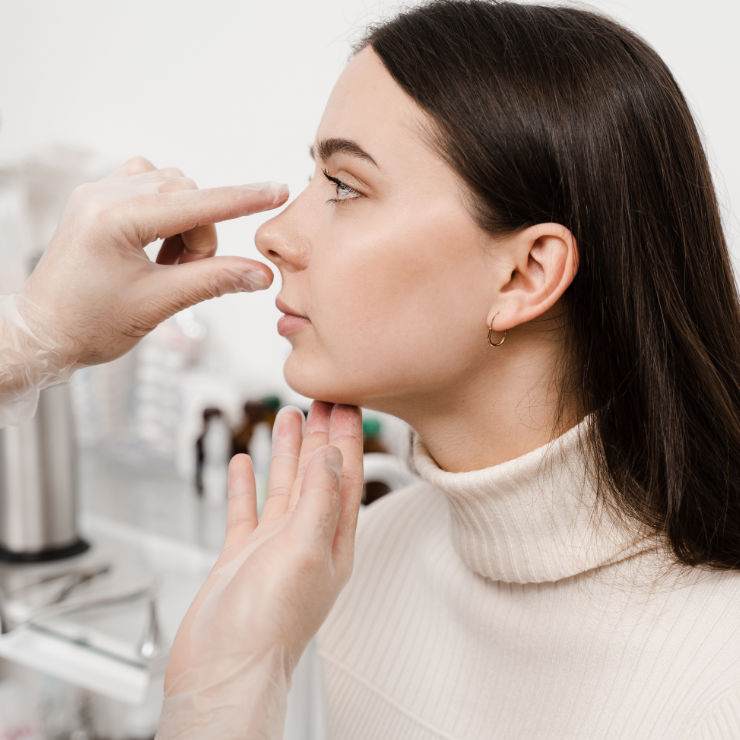 Person in a white sweater undergoing a nose examination by a gloved professional in an office.