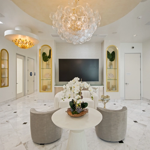 Modern room with marble floor, two beige chairs, round white table, and large TV. Ornate chandeliers hang from the ceiling, and shelves with decor items flank the TV.