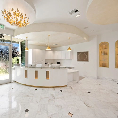 A modern, spacious reception area with white marble floors, gold pendant lights, and a decorative ceiling. The front desk is curved and there are shelves with lights on the right wall.