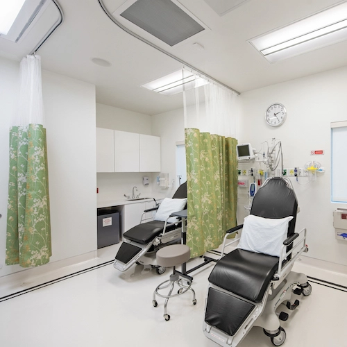 A clean medical room with two treatment chairs, green privacy curtains, wall-mounted medical equipment, and bright overhead lights.