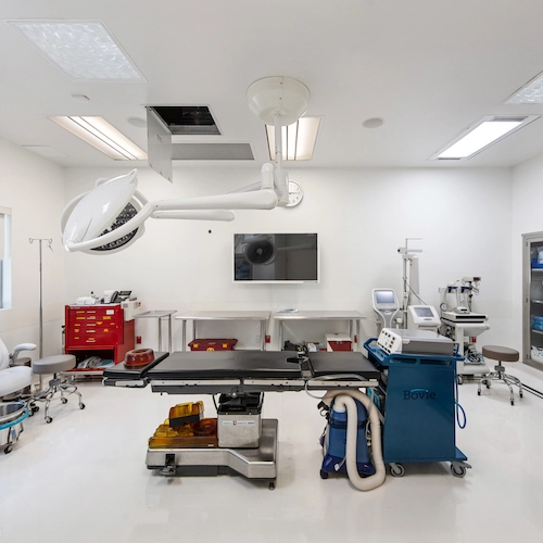 Modern operating room with surgical table, overhead light, medical equipment, and monitors. Walls and floor are white, with various machinery and tools neatly arranged.