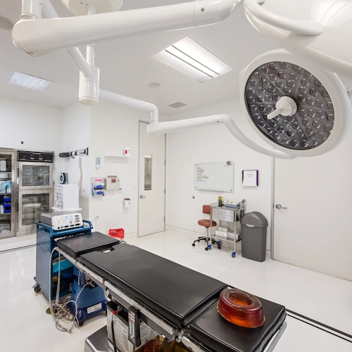 A clean, modern operating room featuring a surgical table, overhead lights, medical equipment, and storage cabinets.