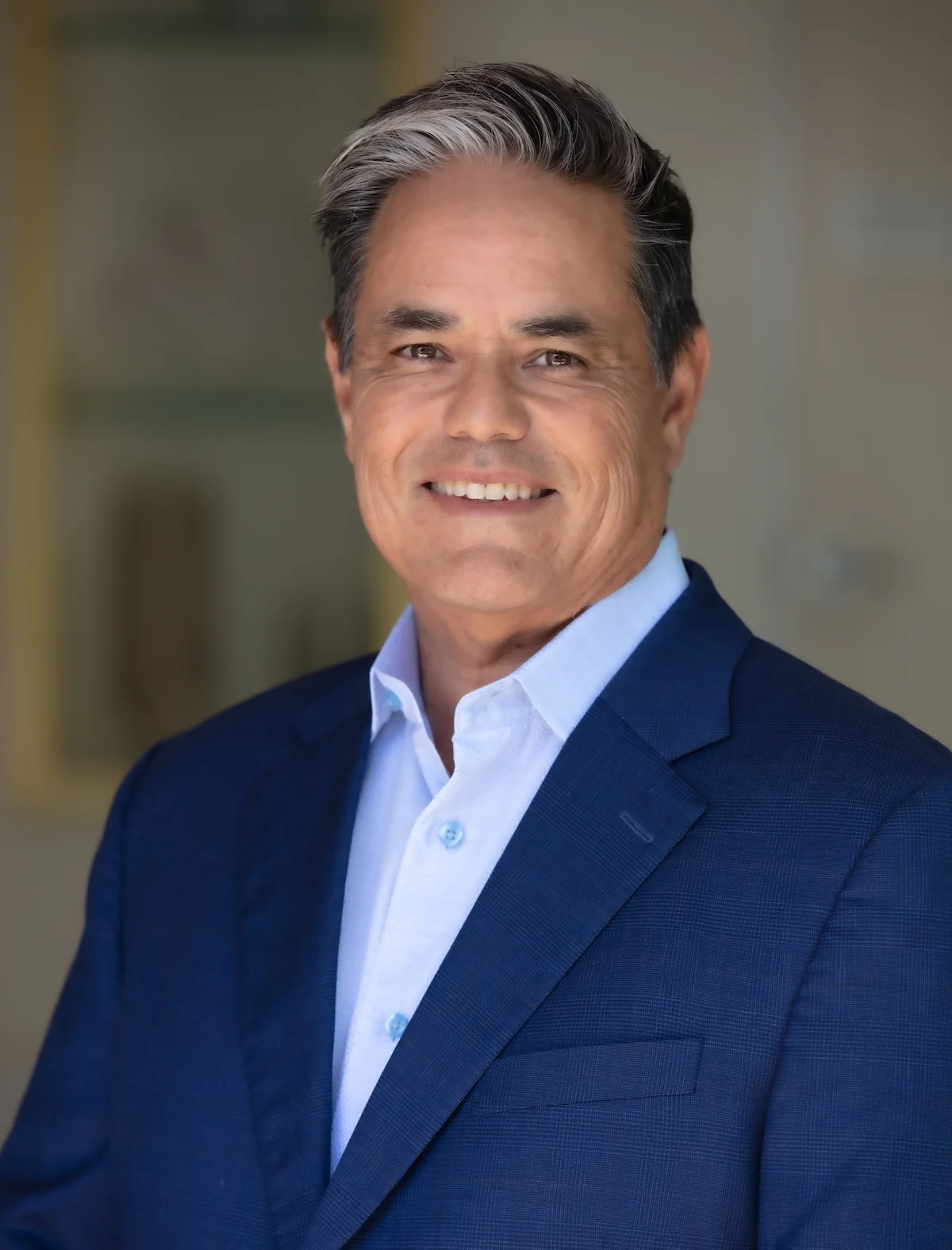 Man in a blue suit jacket and white shirt smiling in a professional portrait. Background is softly blurred.