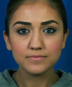 A person with dark hair and makeup looks directly at the camera against a blue background.