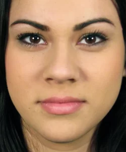 A close-up of a person's face with straight black hair, neutral expression, and pink lips. The background is blurred.