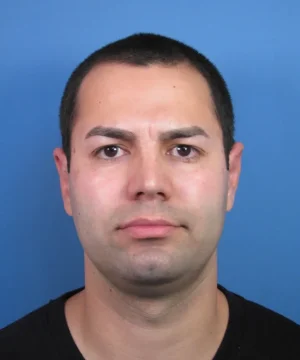 A man with short dark hair and a neutral expression against a blue background. He is wearing a black shirt.