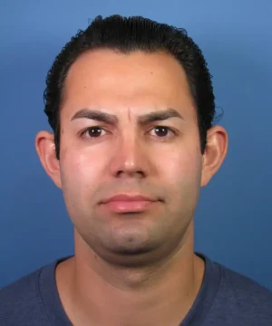 A person with short dark hair against a blue background, wearing a dark-colored shirt, looking directly at the camera.