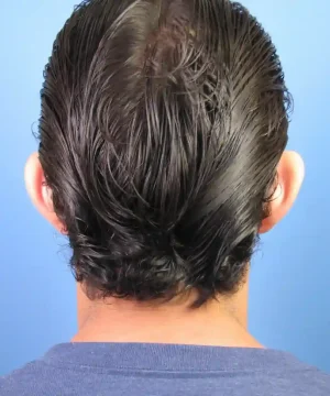 Back of a person's head with wet, slicked-back black hair, wearing a blue shirt, against a plain blue background.