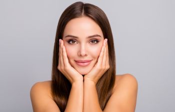 Woman with long brown hair touches her face with both hands against a gray background.