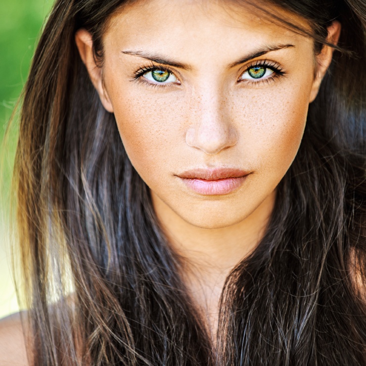 A person with long brown hair and green eyes gazes directly at the camera. The background is blurred and green.