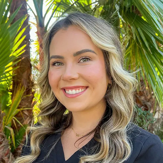 A woman with long, wavy hair smiles in front of palm trees, wearing a black top.