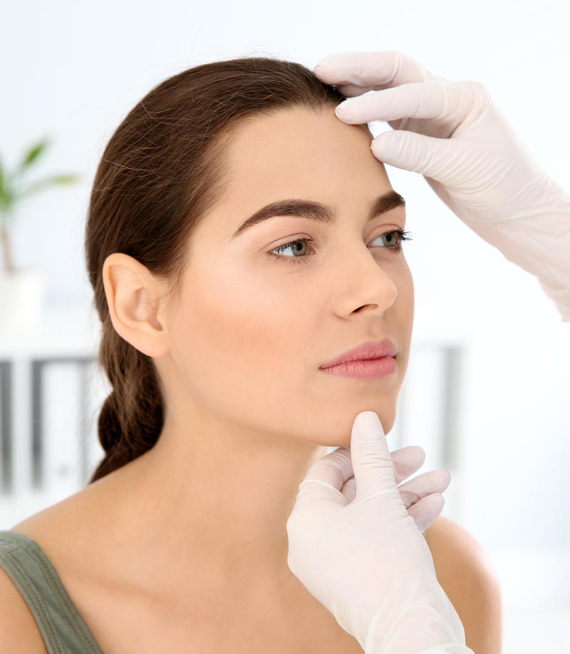 A person with dark hair is examined by gloved hands, touching their forehead and chin, in a clinical setting.