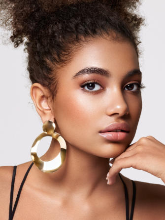 A person with curly hair is wearing large gold hoop earrings and a black strap top, posing with their hand touching their chin against a neutral background.